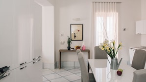 A modern kitchen with white cabinets, a dining table, and chairs. There is a framed picture on the wall, a potted plant, and a vase with flowers on the table. - B&B L'Ambasciata (Gaeta)