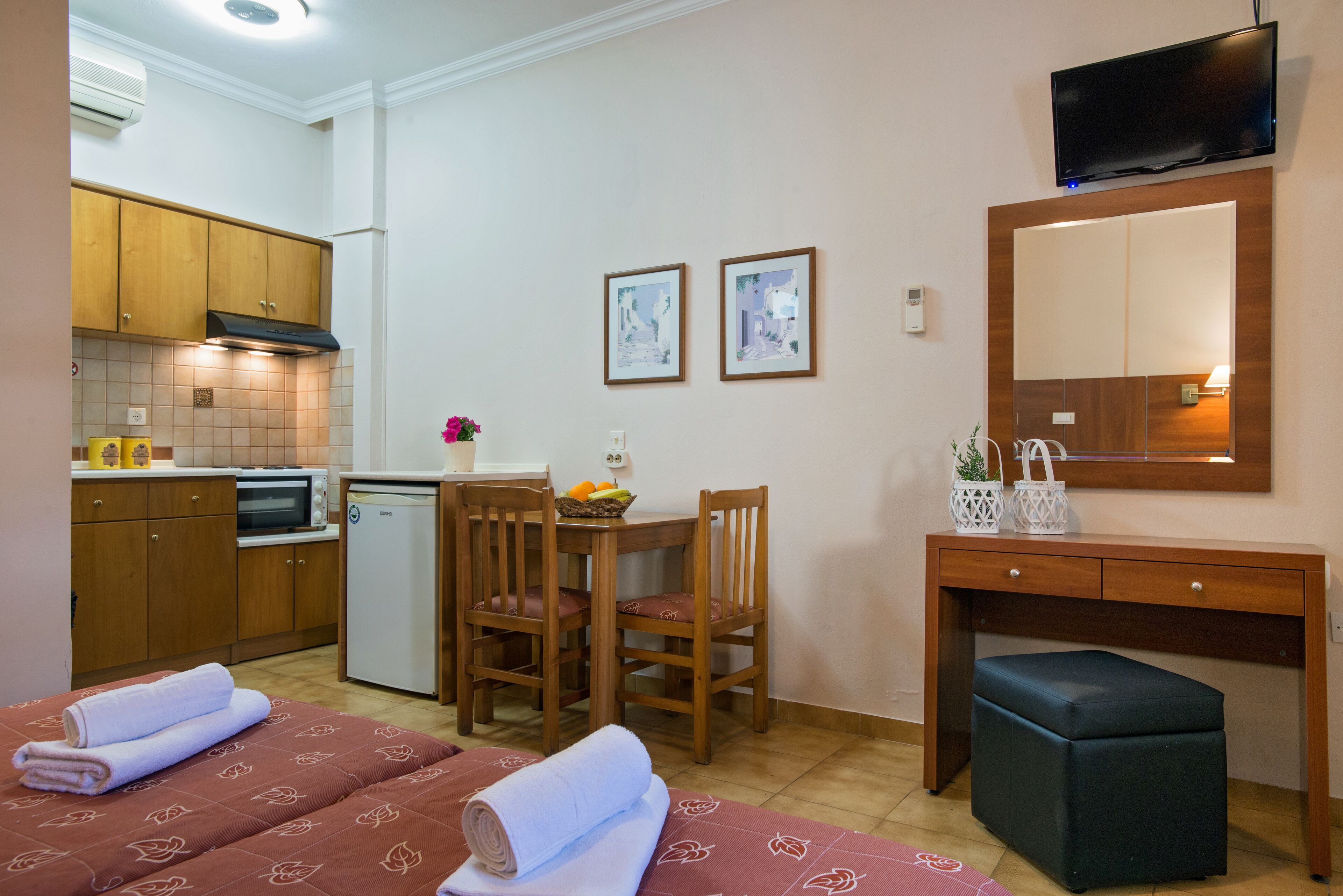 A compact hotel room with a kitchenette, a dining area, a bed with towels, a desk, and a TV.