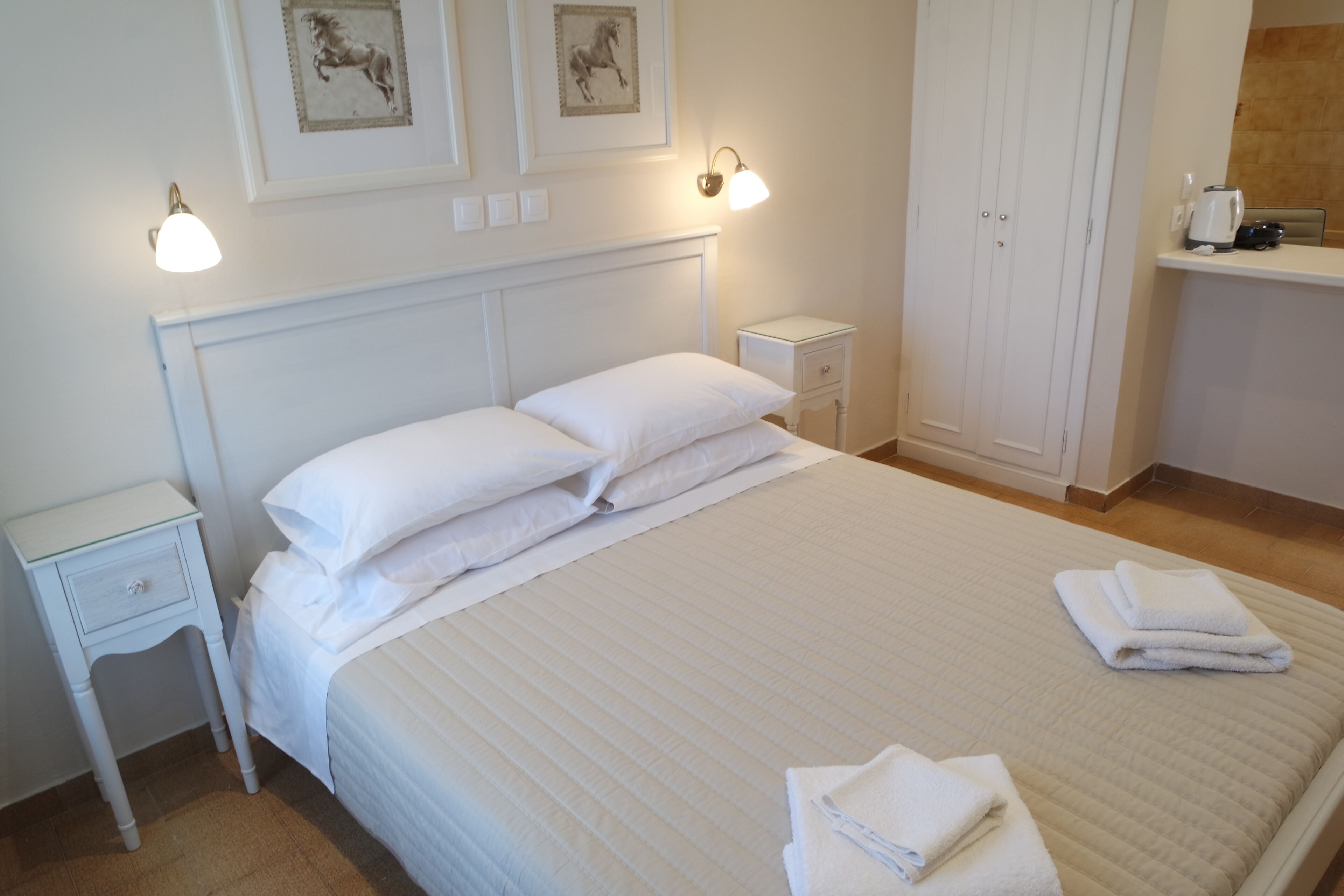 Een netjes opgemaakt bed met witte lakens en kussens, een nachtkastje met een lamp en een kledingkast in een hotelkamer.