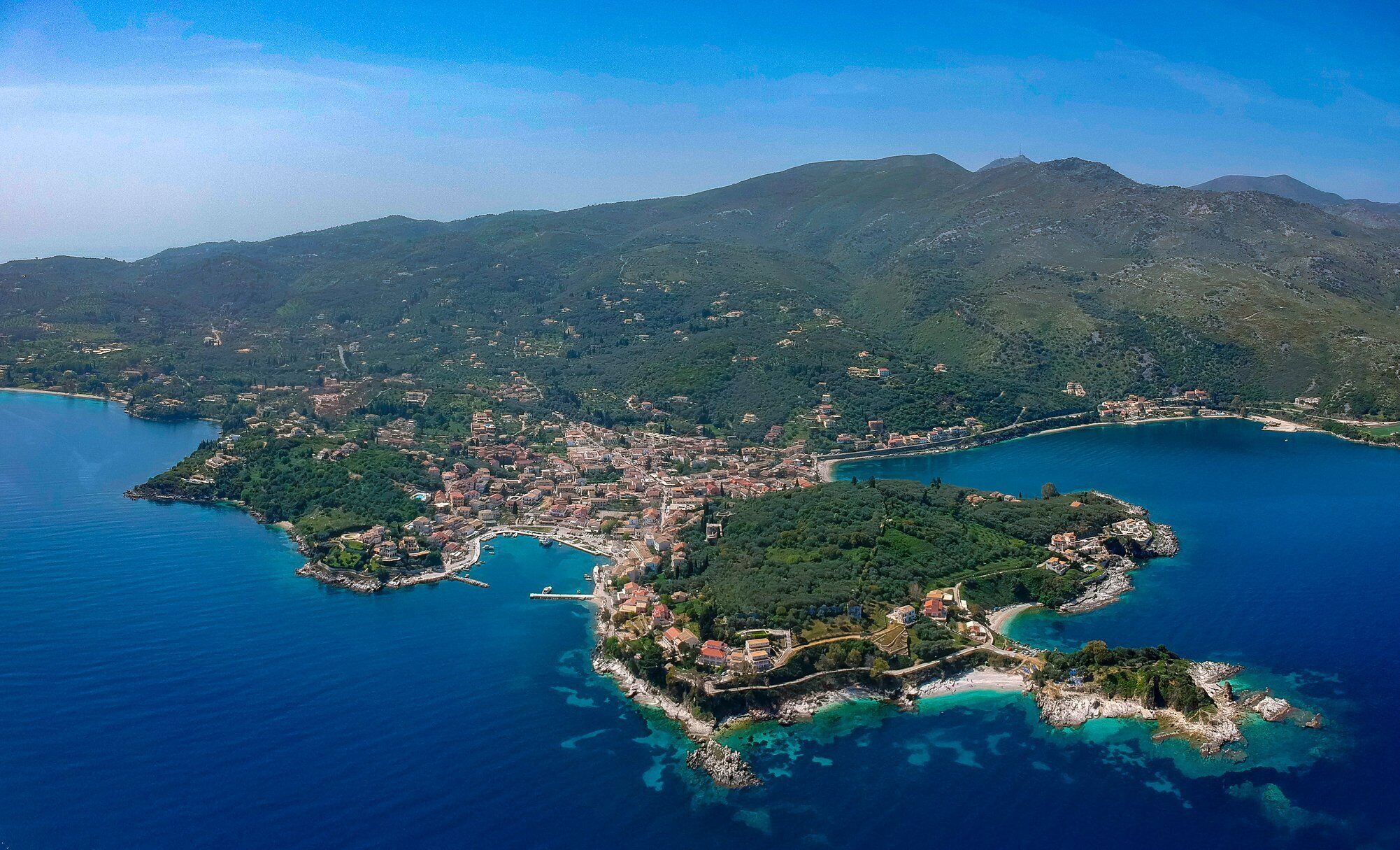 An aerial view of a coastal town with a central island, surrounded by mountains and clear blue waters.
