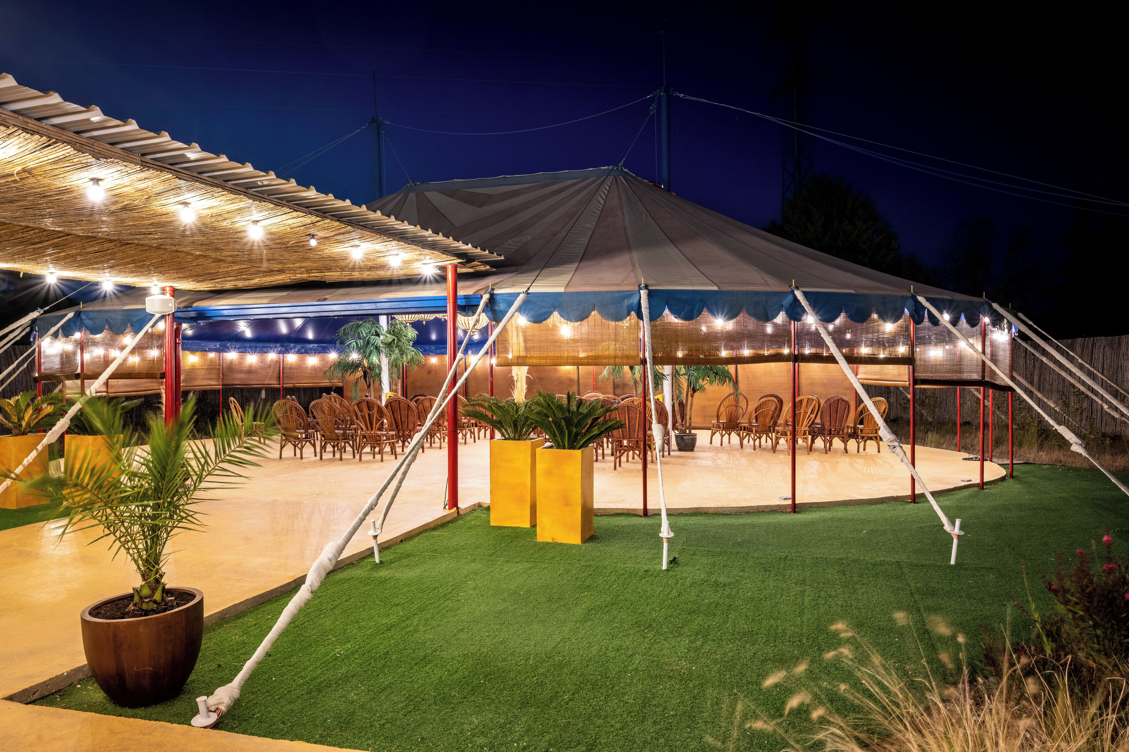 A covered outdoor seating area with tables and chairs, surrounded by artificial grass and potted plants.
