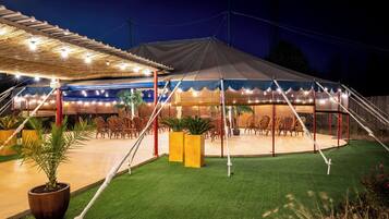 A covered outdoor seating area with tables and chairs, surrounded by artificial grass and potted plants.