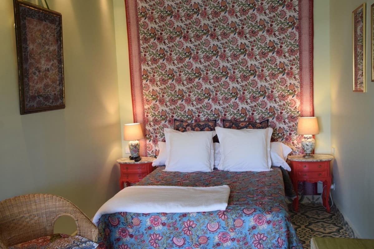 A bed with a floral headboard, two bedside tables with lamps, a wicker chair, and a framed picture on the wall.