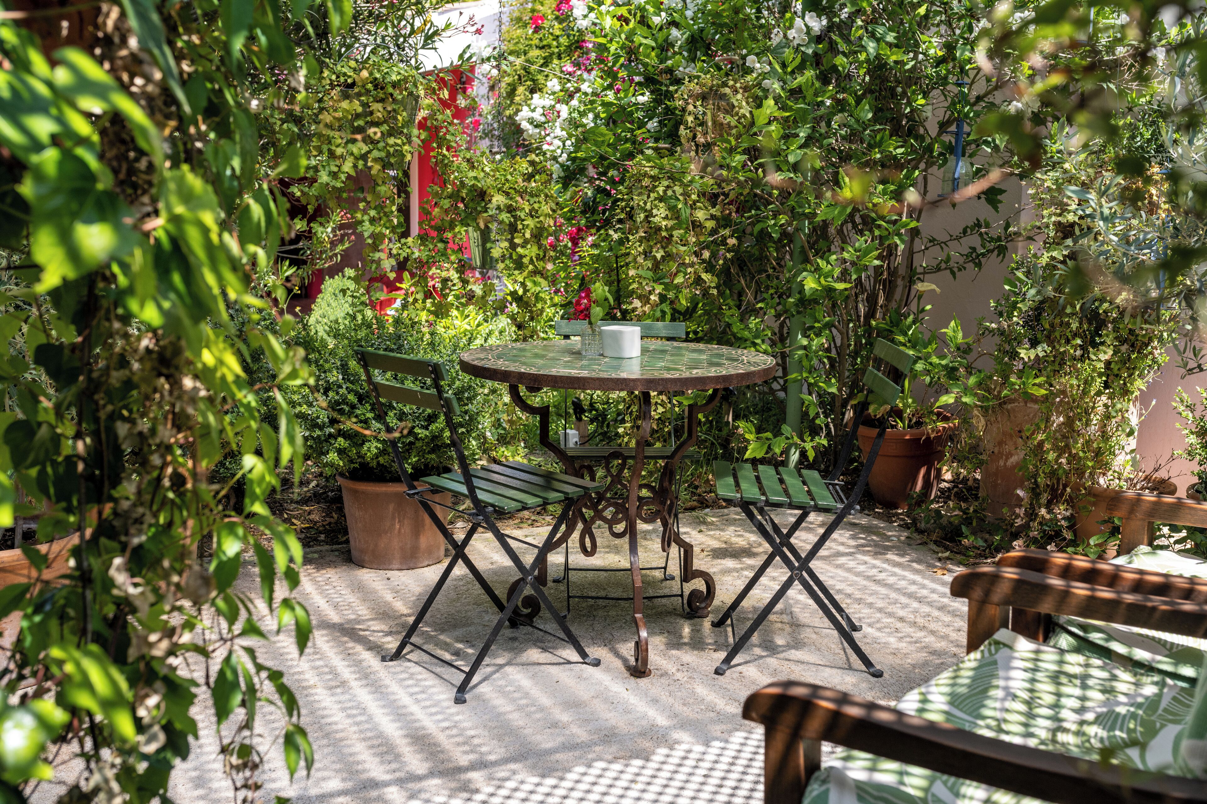 Una mesa metálica redonda con un banco verde, rodeada de plantas en macetas y un suelo de hormigón.