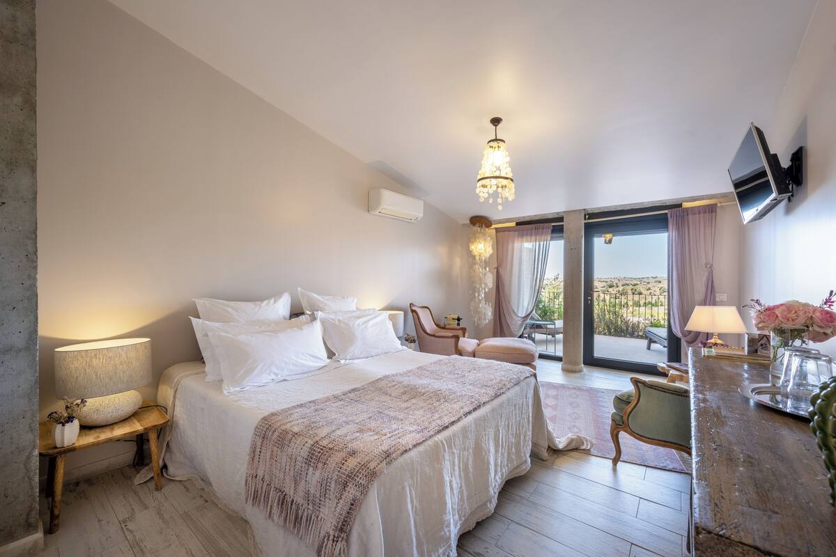 A bedroom with a large bed, a desk, a chair, and a balcony with a view.