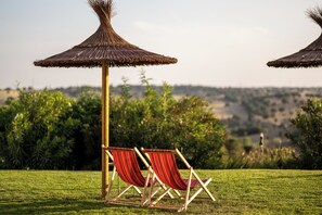 Two red striped lounge chairs under a thatched umbrella in a grassy area with a view of a hilly landscape. - Finca Aal Cachucho Hotel Rural (San Agustin del Guadalix)