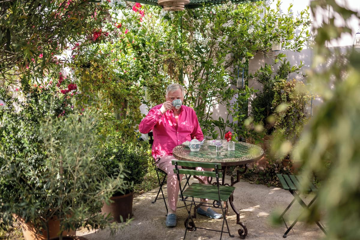 A person sitting at a round table with a teapot and cups, surrounded by lush greenery and potted plants.