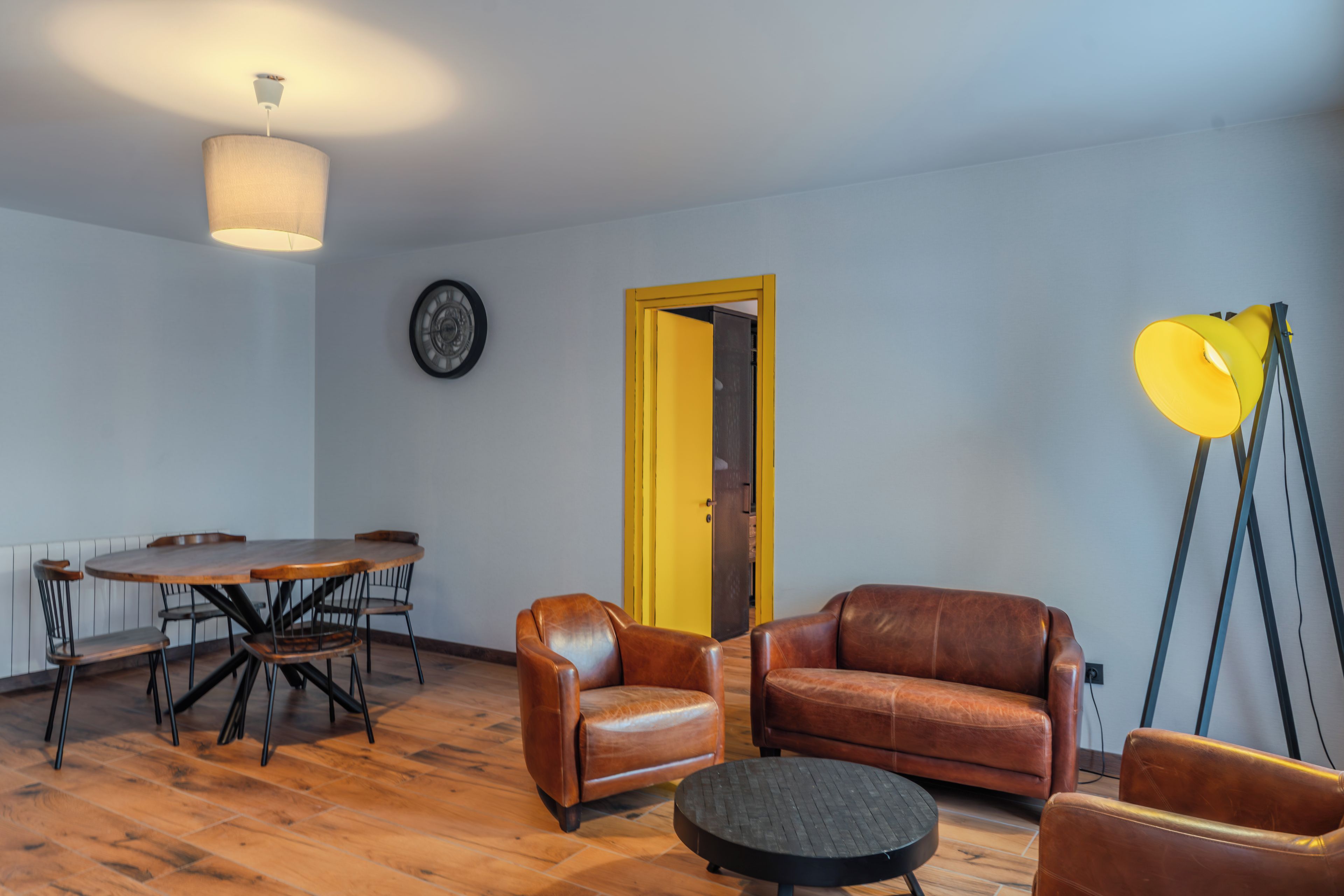 A room with a dining table, two leather chairs, a sofa, a floor lamp, and a door.