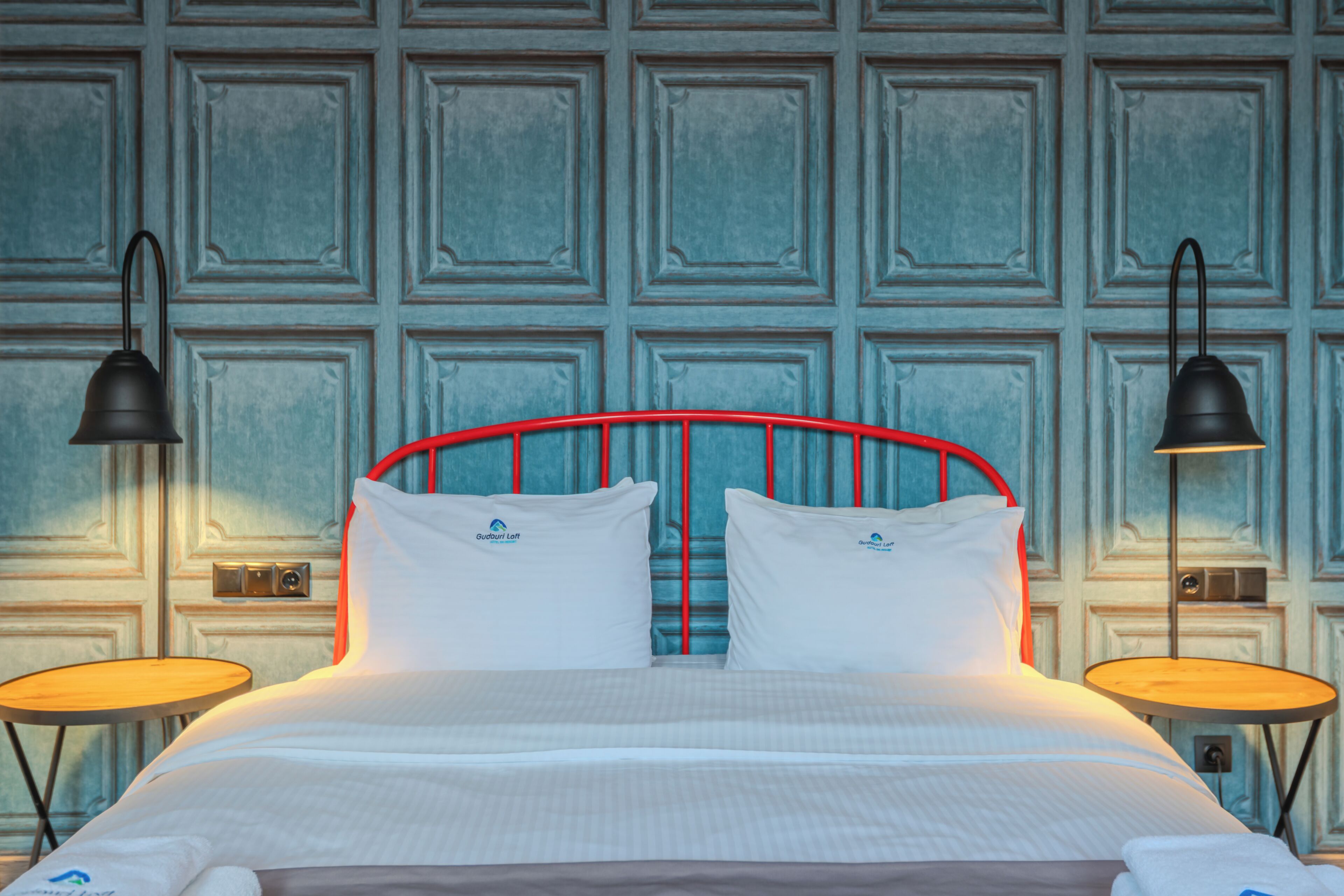 A hotel room with a red metal headboard, two white pillows, a bed with a white bedspread, two bedside tables with lamps, and a blue paneled wall.