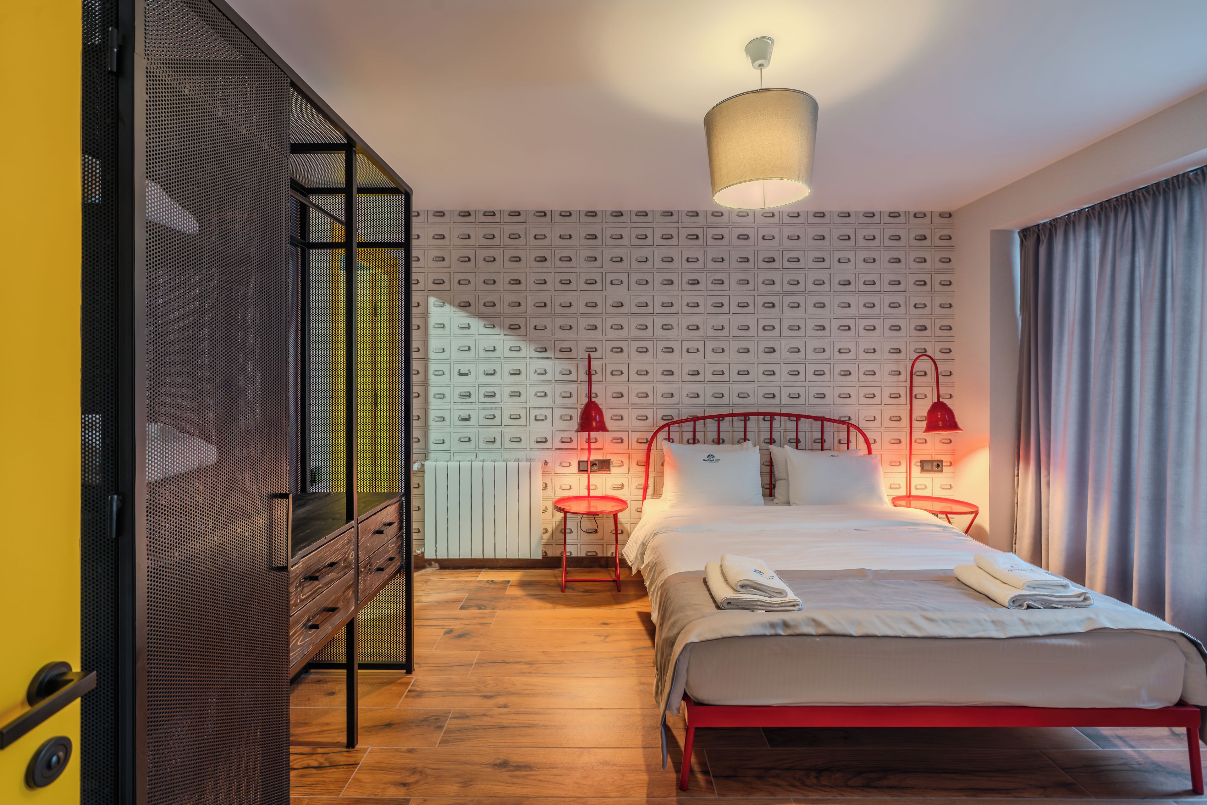 Habitación de hotel con cama, lámpara roja, mesita de noche roja, radiador blanco y pared con estampado.