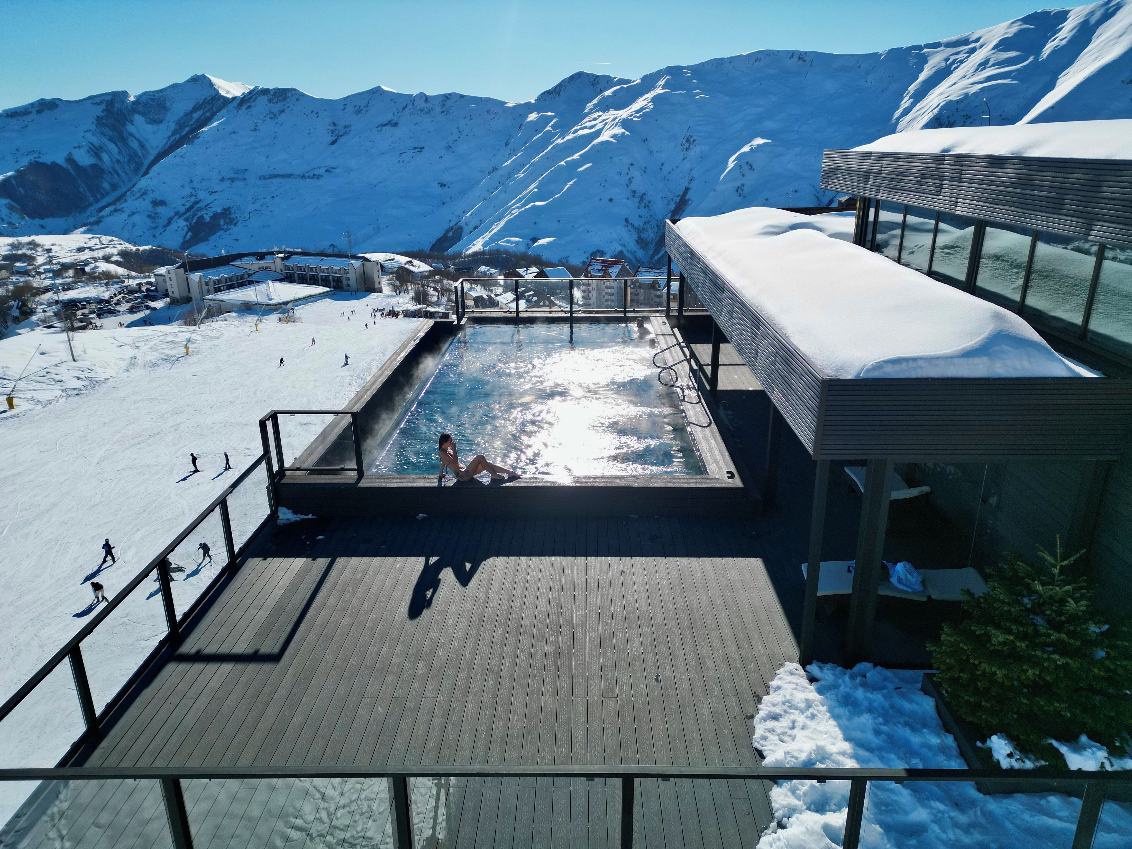 Area piscina con vista su montagne innevate, una pista da sci e un edificio con terrazza coperta.