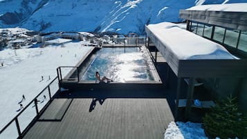 A pool area with a view of snow-covered mountains, a ski slope, and a building with a covered deck.