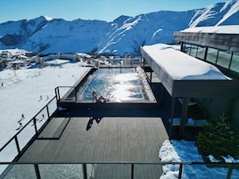 Zona de piscina con vistas a montañas nevadas, una pista de esquí y un edificio con terraza cubierta.