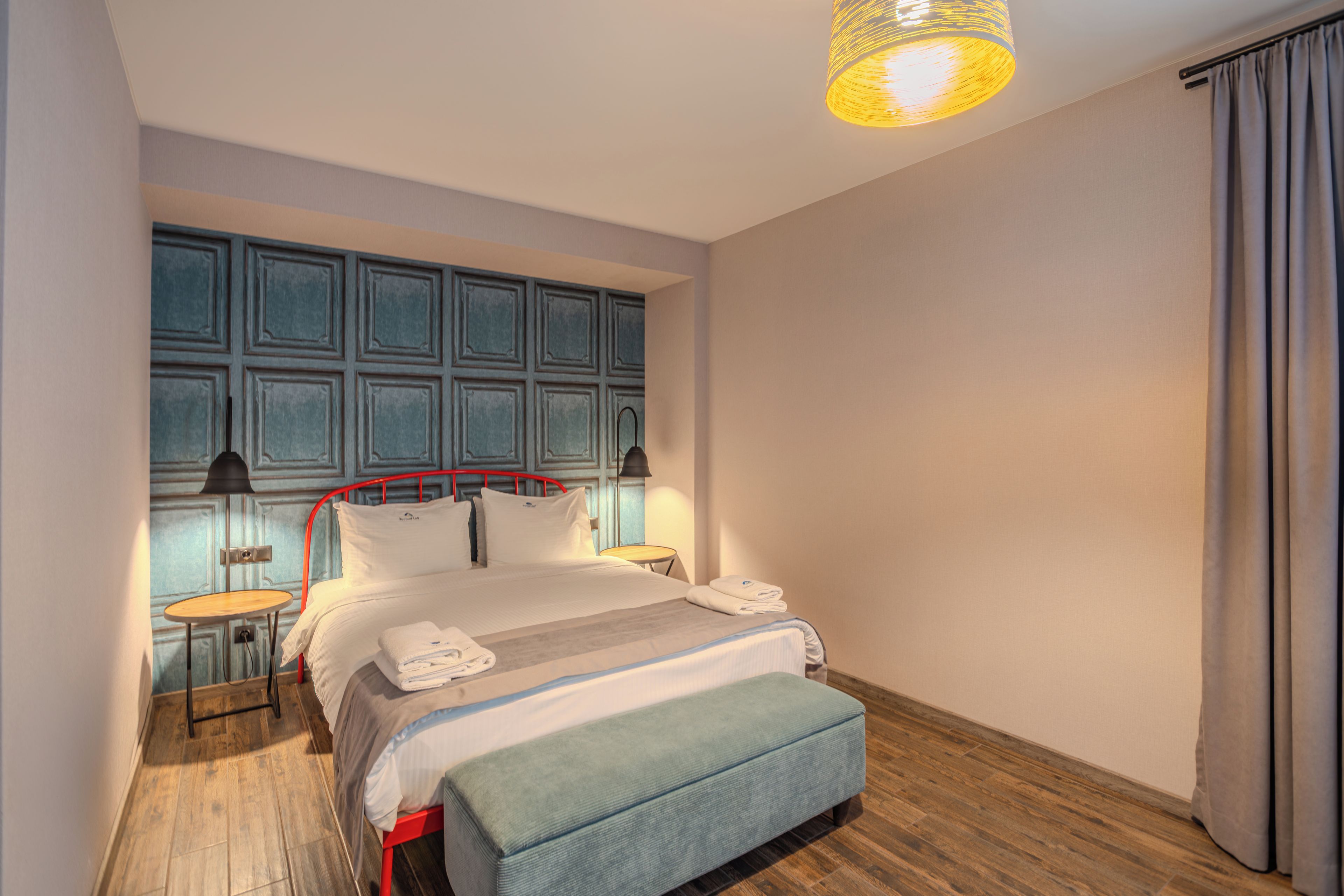 A hotel room with a bed, bedside tables, a lamp, a bench, a window with curtains, and a wall with a panel design.