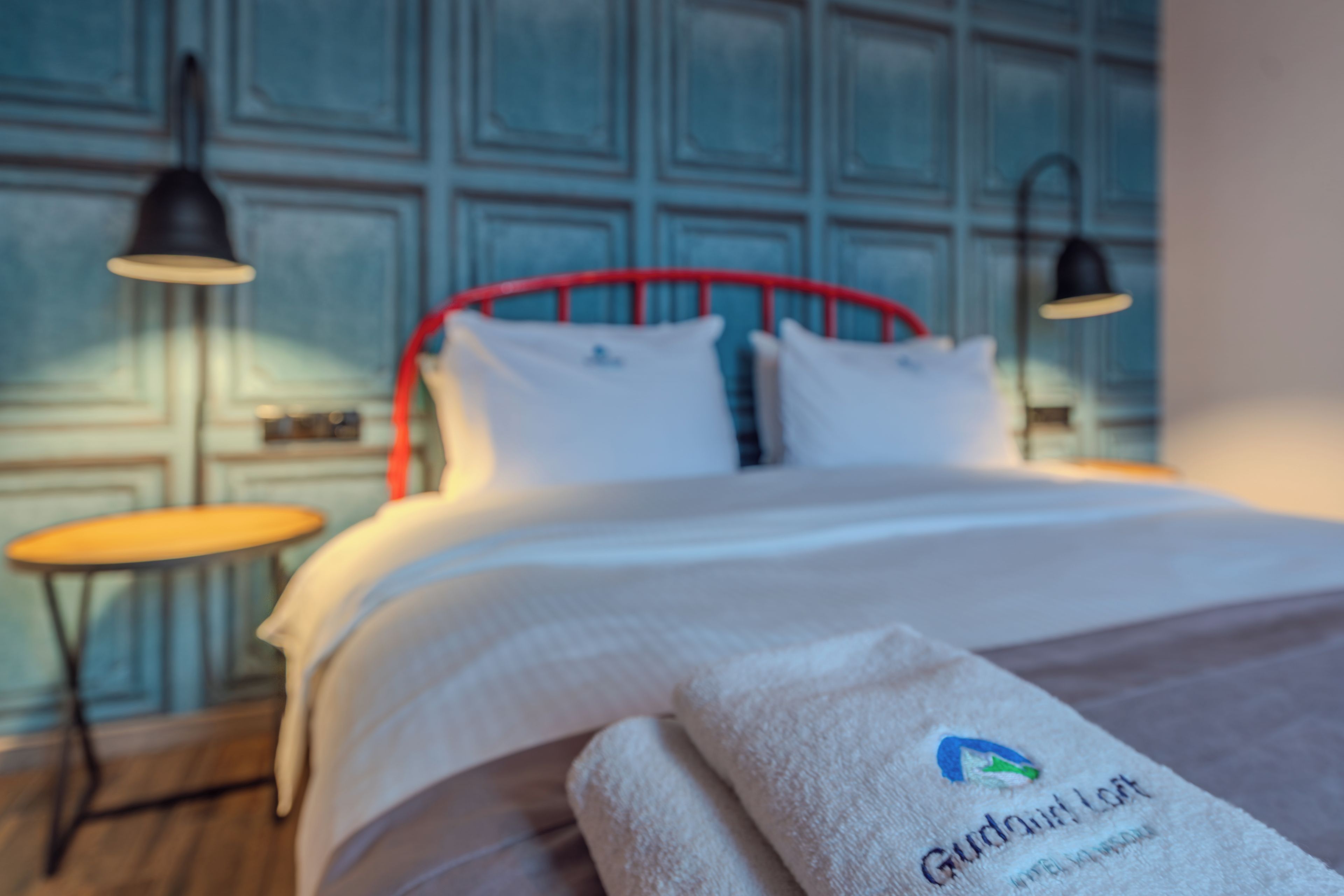 A neatly made bed with a red metal frame, white bedding, and a towel with a logo on it. There are two bedside tables with lamps, one on each side of the bed.