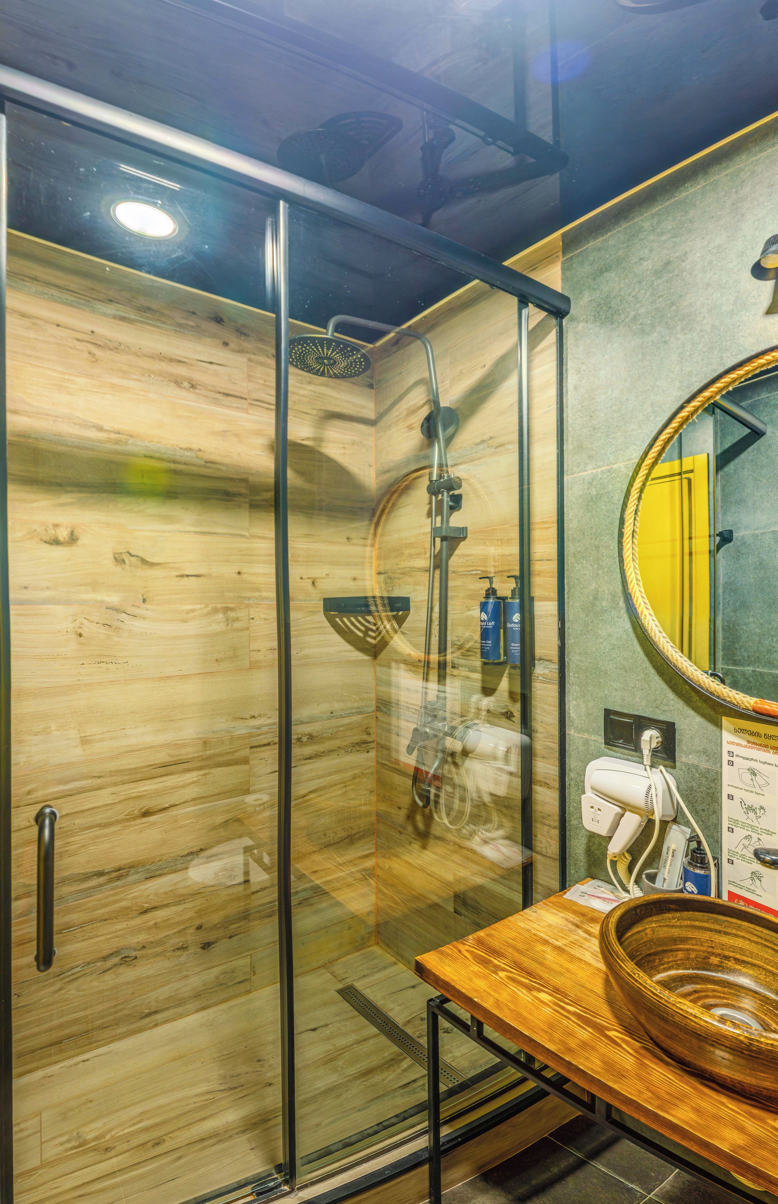 Una ducha con cerramiento de vidrio, interior de madera, un tocador de madera con lavabo y un espejo con marco amarillo.