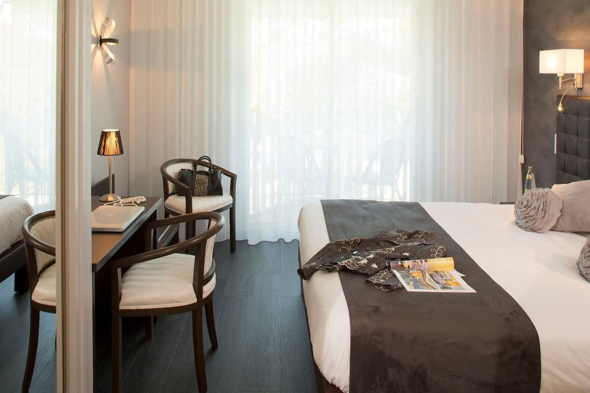 A hotel room with a bed, a desk, two chairs, a lamp, and a window with sheer curtains.