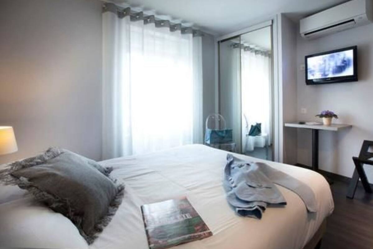 A modern hotel room with a large bed, a flat-screen TV mounted on the wall, a small table with a plant, and a view of the city through the window.