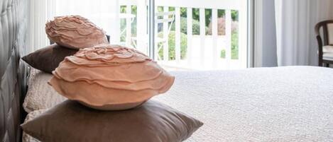 A bed with a textured white cover, two brown pillows, and a view of a white balcony with greenery outside.