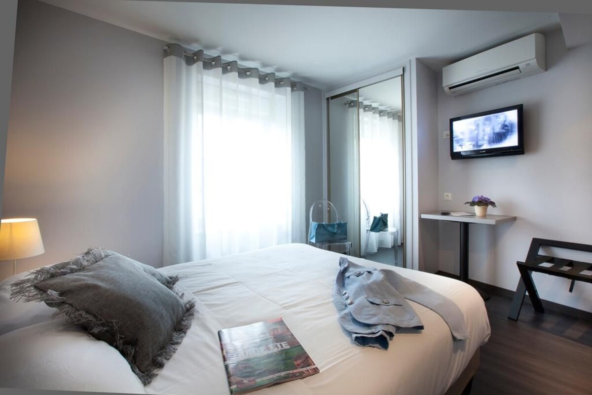 Quarto de hotel com cama, televisão, pequena mesa com uma planta e um banco.