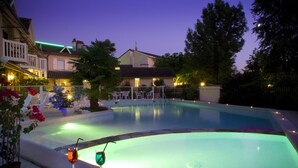 A swimming pool at dusk with a table and drinks, surrounded by buildings and trees. - Hotel Le Relais de Farrou (Saint-Remy)