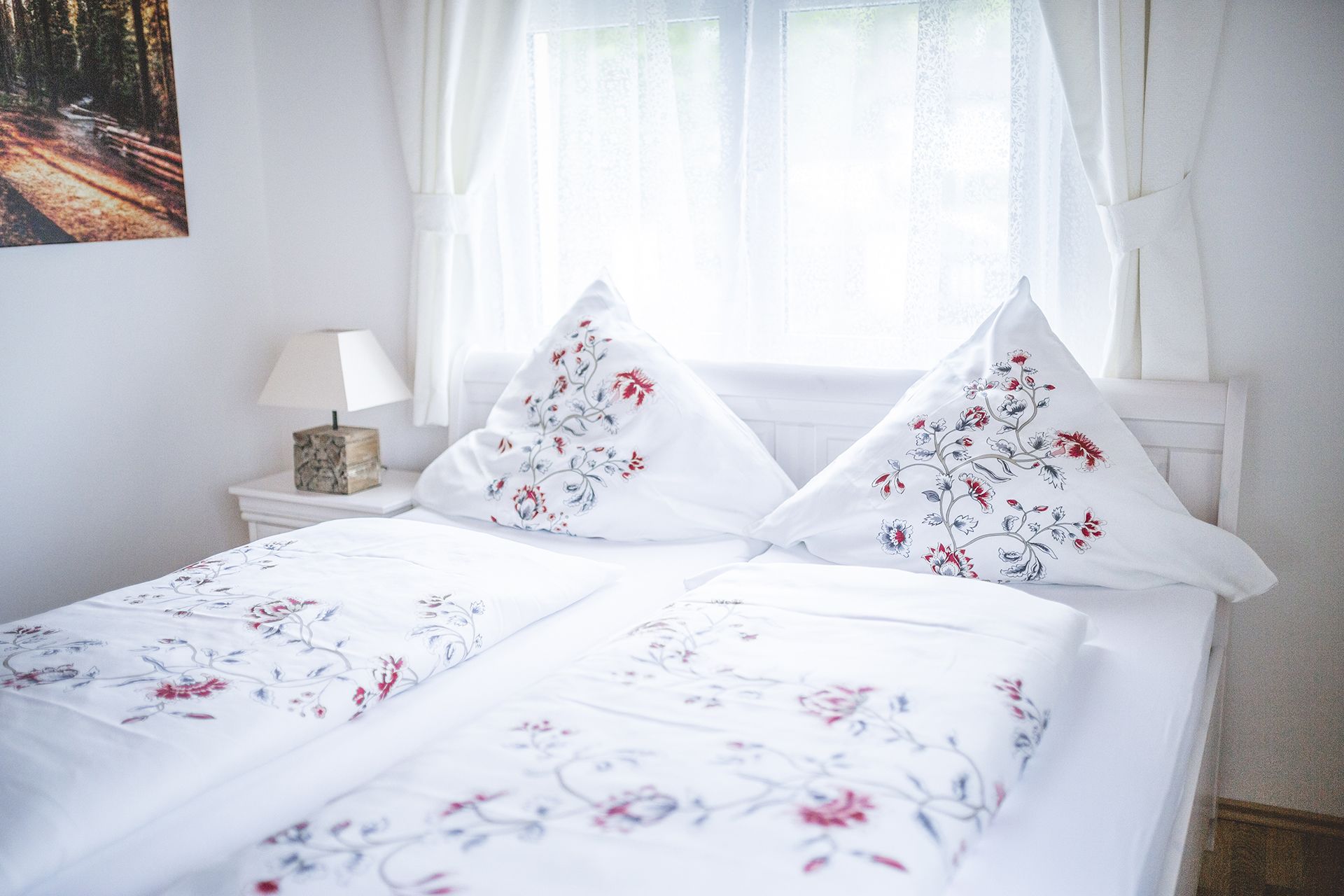 A neatly made bed with floral bedding, a bedside table with a lamp, and a window with sheer curtains.