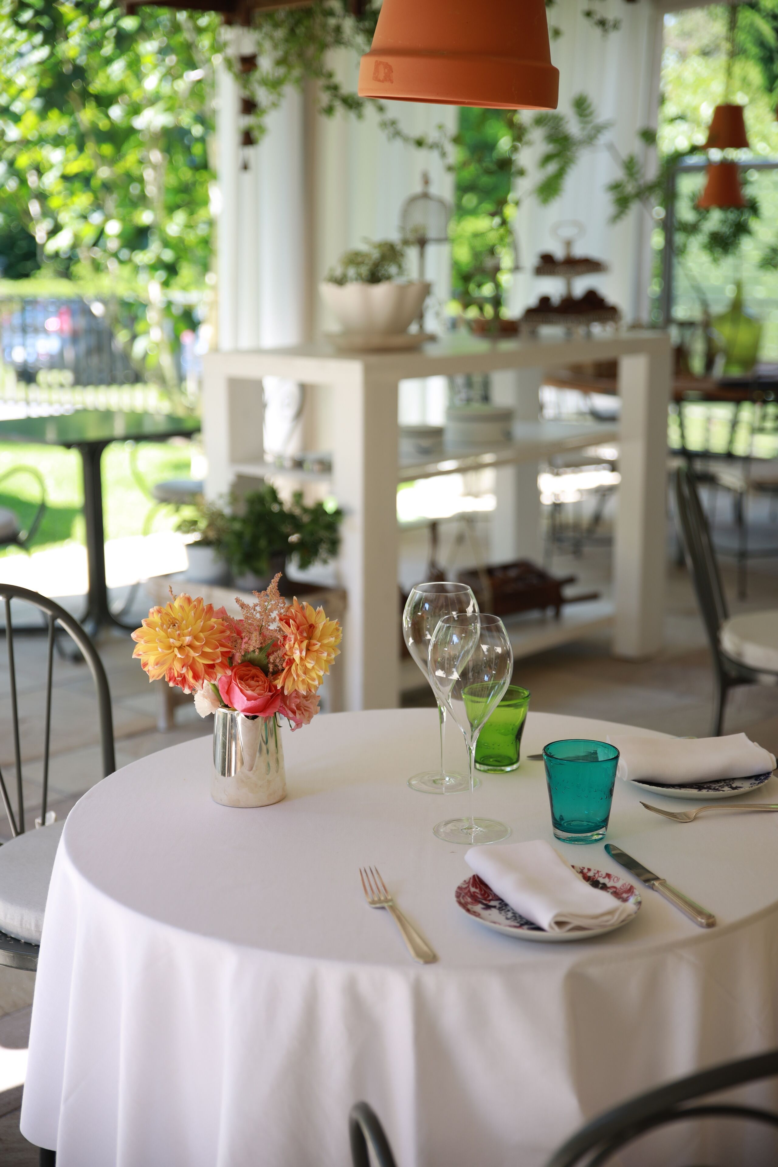 A round table with a white tablecloth, a vase with flowers, wine glasses, and a green glass.