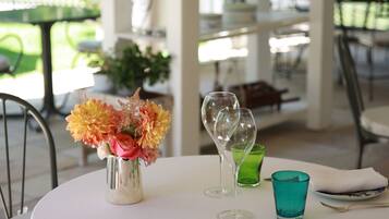 Una mesa redonda con mantel blanco, un florero con flores, copas de vino y un vaso verde.