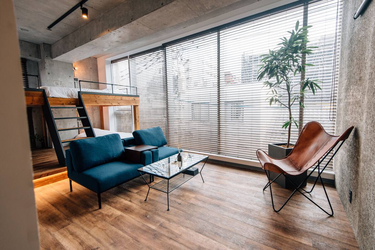 Un espacio habitable moderno con cama tipo altillo, sofá azul, sillón de cuero y un amplio ventanal con persianas.