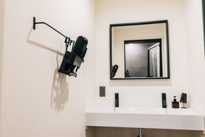 Une salle de bain avec un lavabo blanc, un miroir et un sèche-cheveux noir fixé au mur.