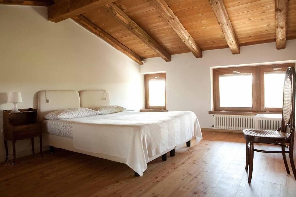 A bedroom with a wooden ceiling, a bed with white bedding, a chair, and a window.