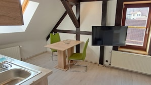 A compact kitchen with a dining area, a TV mounted on the wall, and a window with a view of buildings. - Gästehaus Tröbs (Harztor)