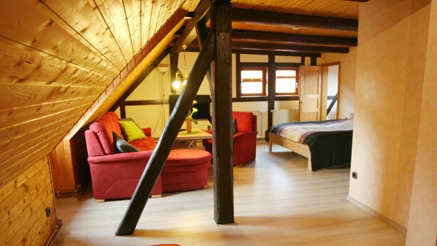 A loft-style bedroom with a sloped ceiling, wooden beams, a bed, a red chair, and a small table with a plant. - Gästehaus Tröbs (Harztor)