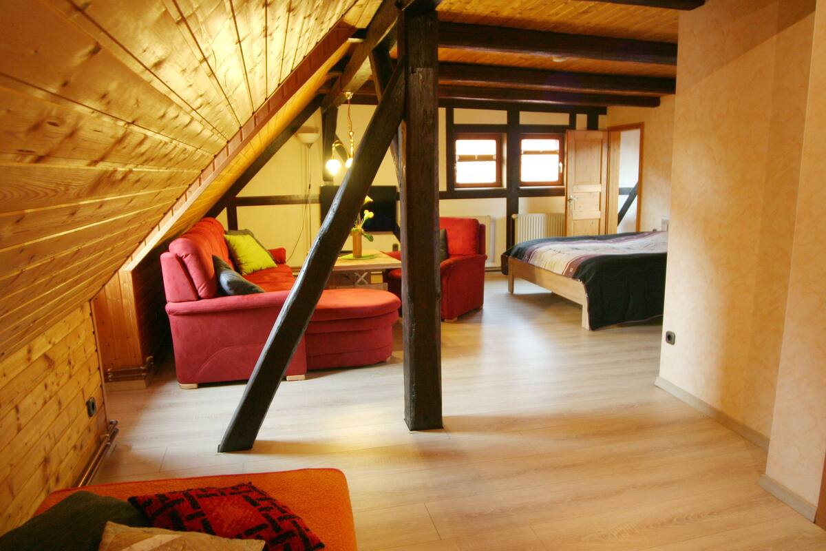 Ein Schlafzimmer im Loft-Stil mit schrägem Dach, Holzbalken, einem Bett, einem roten Sessel und einem kleinen Tisch mit Pflanze.