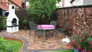 A patio with a brick floor, a round table with chairs, a pizza oven, and a potted plant with pink flowers. - Gästehaus Tröbs (Harztor)