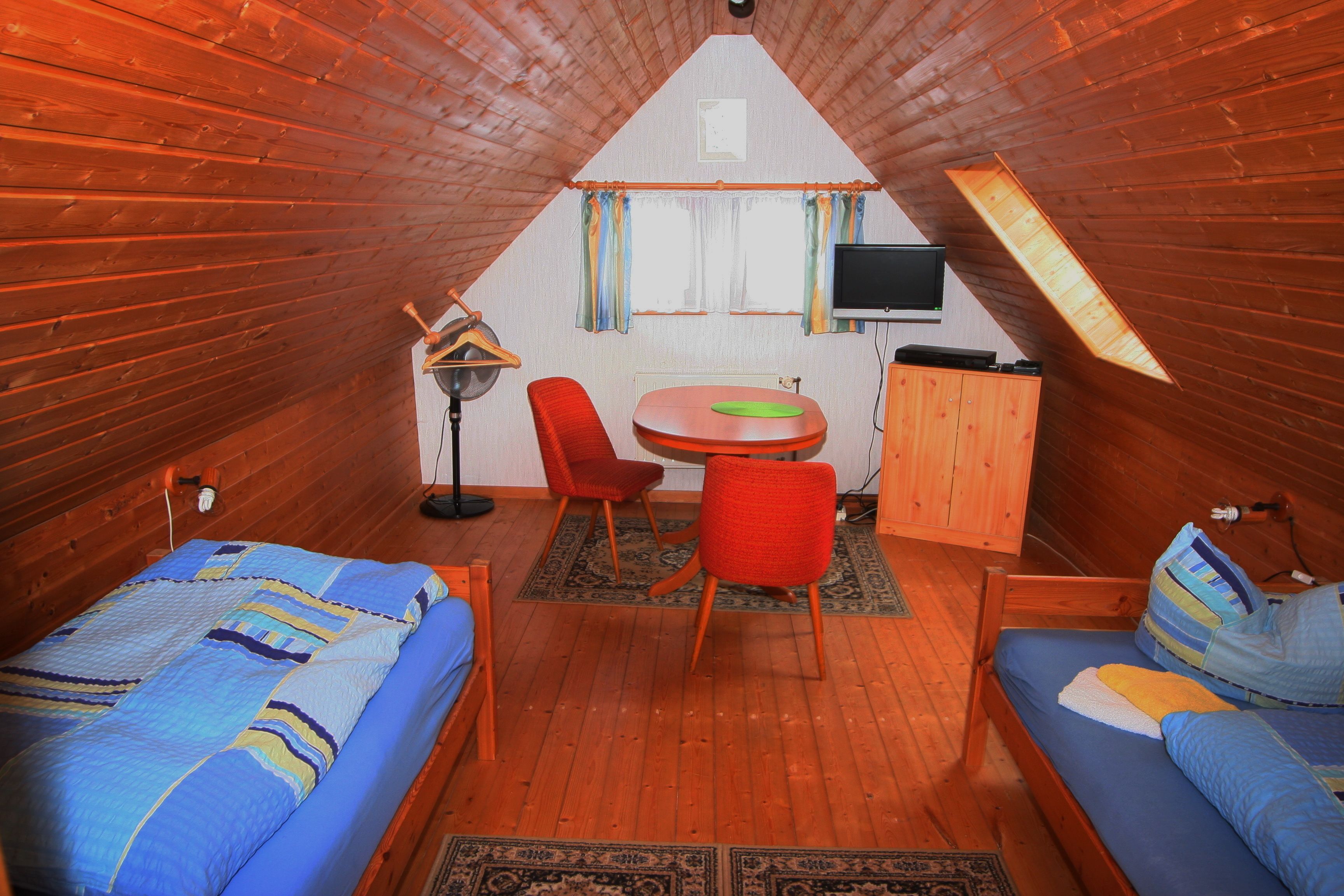 A cozy attic room with a wooden floor, two beds, a small dining table with chairs, a TV mounted on the wall, and a wooden cabinet.