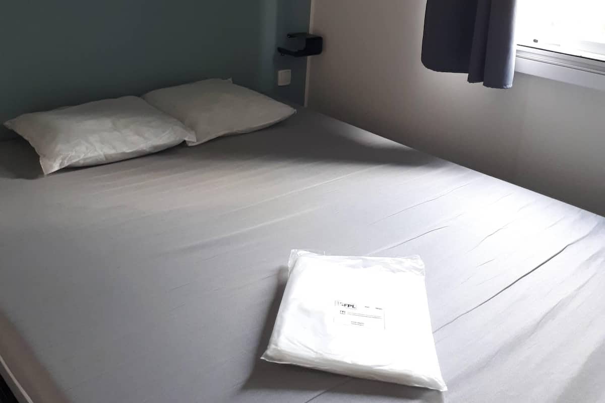 A neatly made bed with white linens, a wooden shelf above it, a bedside table with a lamp, and a window with curtains.