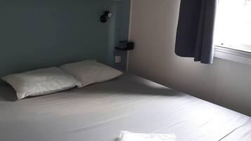 Un lit bien fait, avec du linge de lit blanc, une étagère en bois au-dessus, une table de chevet avec une lampe et une fenêtre avec des rideaux.