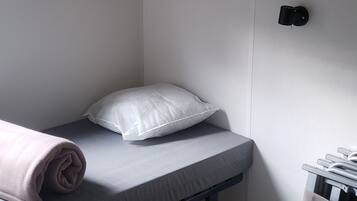 A single bed with a grey mattress, a rolled-up blanket, and a white pillow in a room with wooden flooring and white walls.