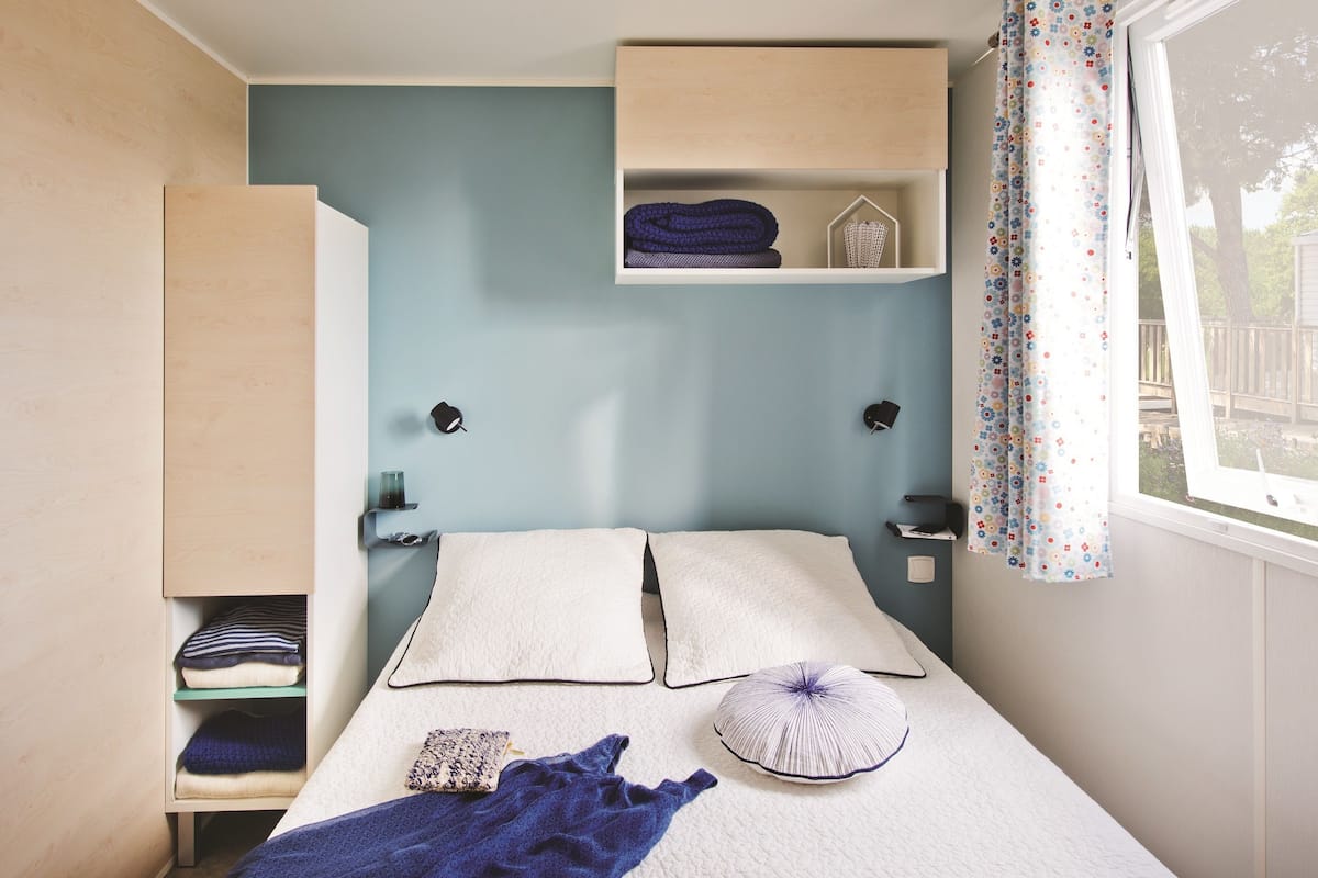 A bedroom with a bed, pillows, a blanket, a shelf with folded clothes, and a window with curtains.