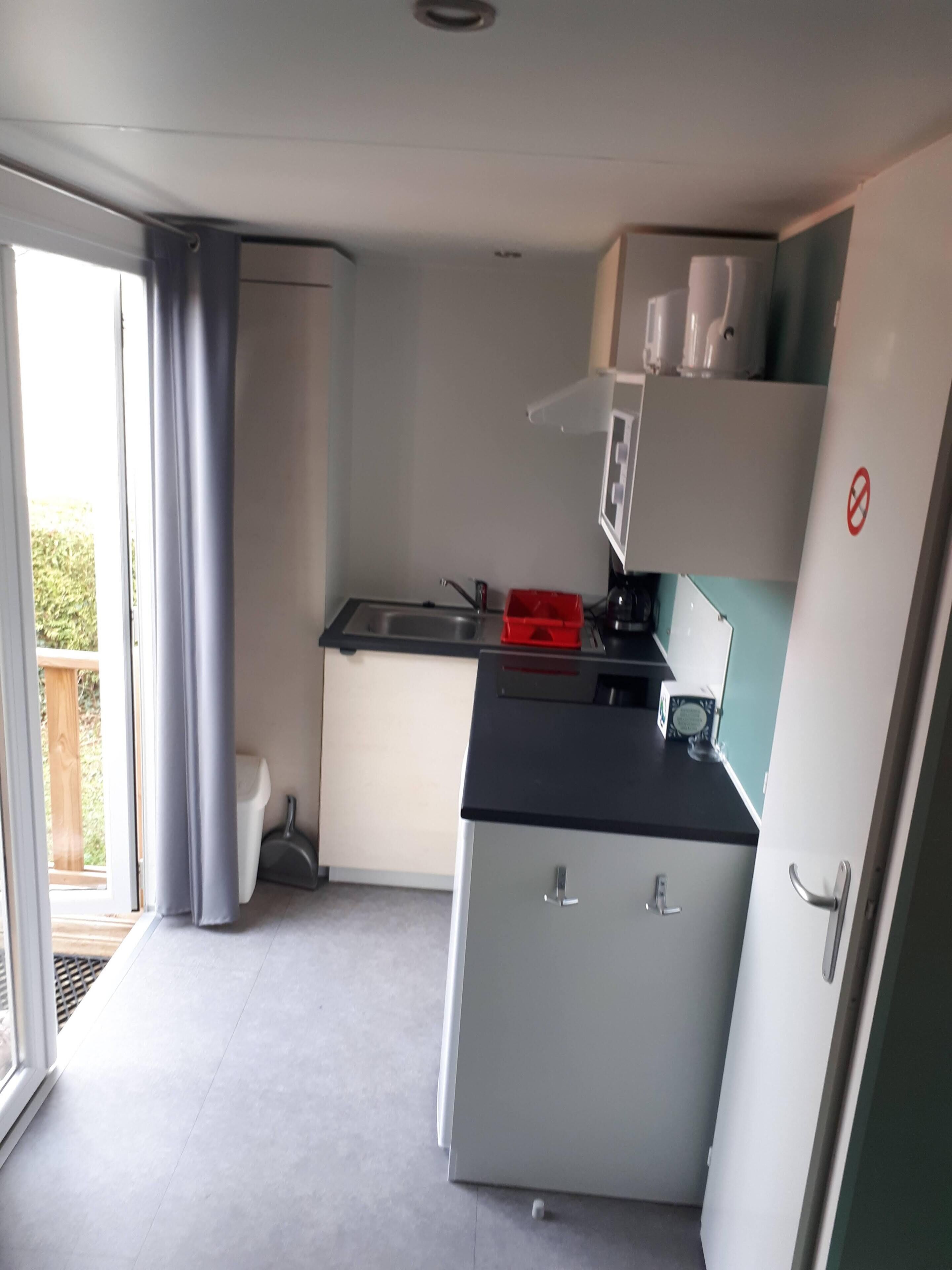A compact kitchen with a sink, a refrigerator, and a microwave. There is a window with curtains, and a door leading to an outdoor area.