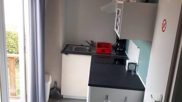 Een kleine keuken met een spoelbak, een koelkast en een magnetron. Er is een raam met gordijnen, en een deur die leidt naar een buitenruimte.