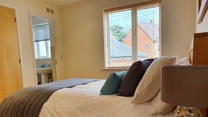 A neatly made bed with a grey blanket, a bedside table with a lamp, and a view of a house through a window with blinds. - Villetts House (Swindon)