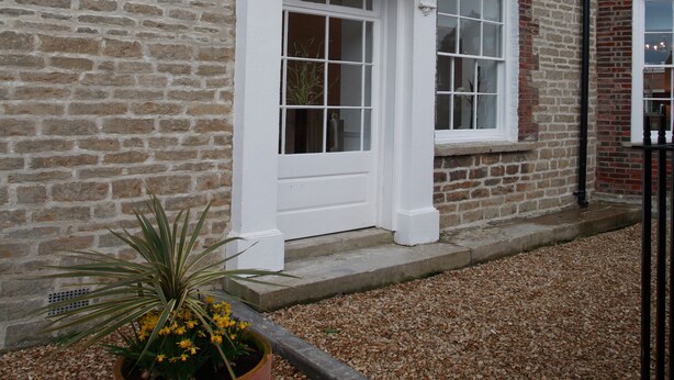 A brick building with a white door and window, a potted plant, and gravel ground. - Villetts House (Swindon)