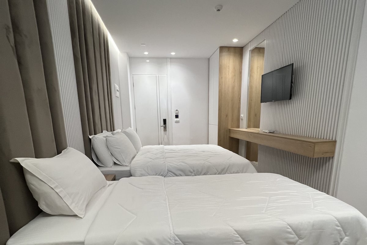 A modern hotel room with two beds, a wooden nightstand, a TV mounted on the wall, and a wooden bench.
