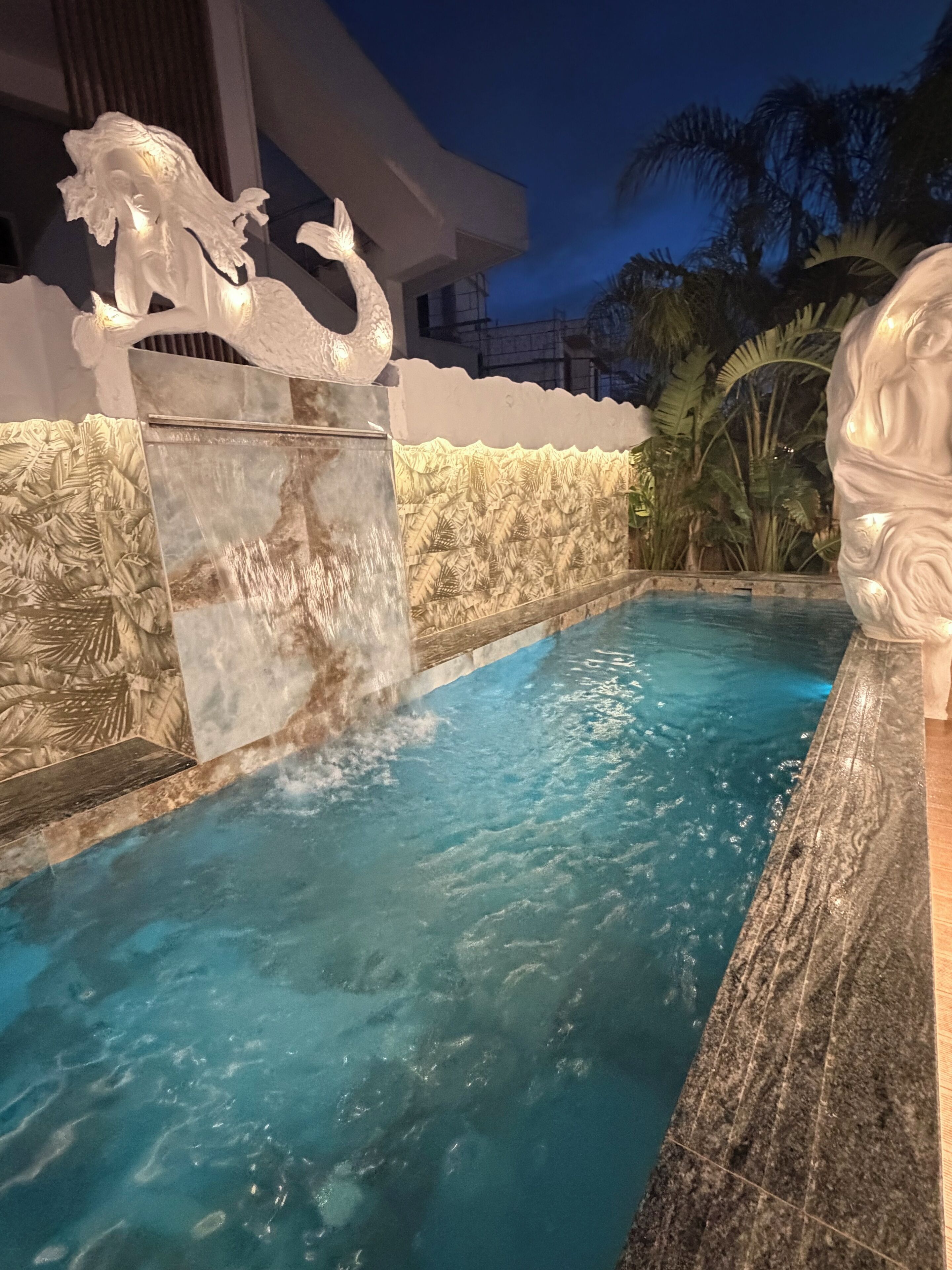 A swimming pool with a waterfall feature, surrounded by a stone wall with carved designs, and a statue on the right.