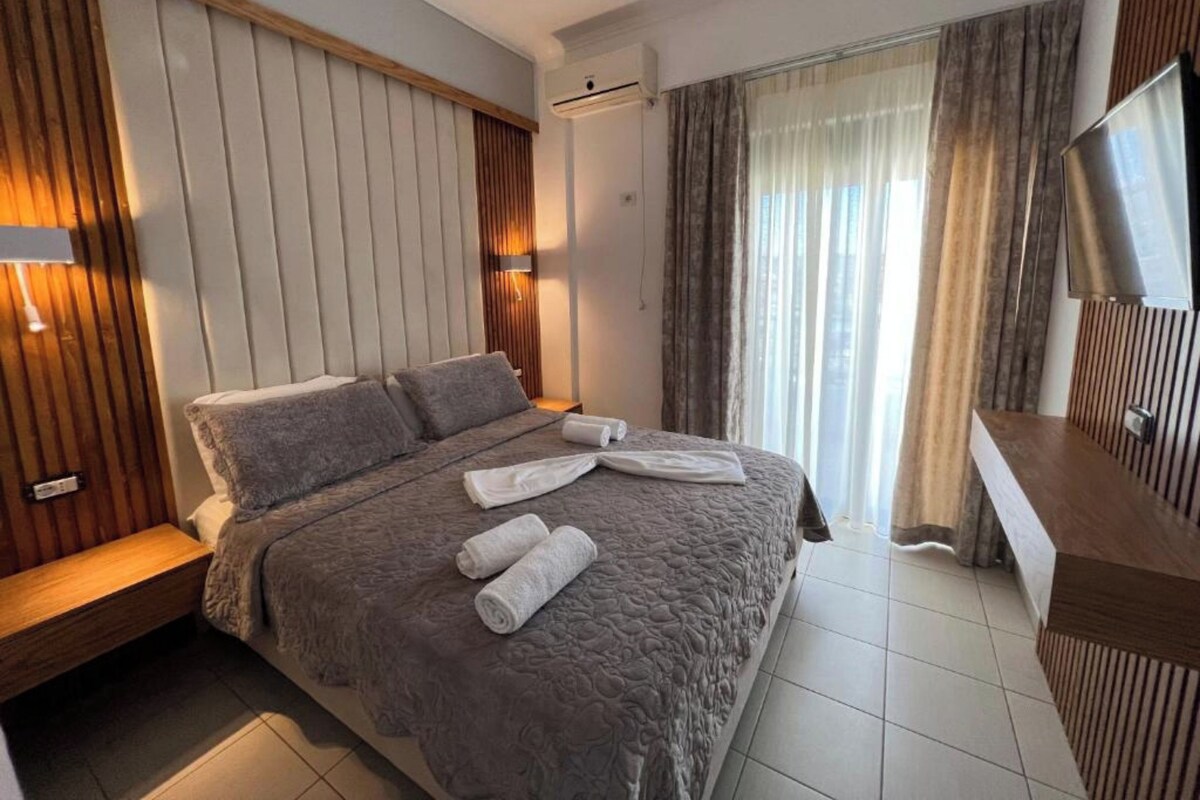 A modern hotel room with a large bed, a wooden headboard, a flat-screen TV, and a window with curtains.