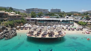 Ein Hotel direkt am Strand mit Poolbereich, mehreren Sonnenschirmen und einer Pier, die ins Wasser führt.