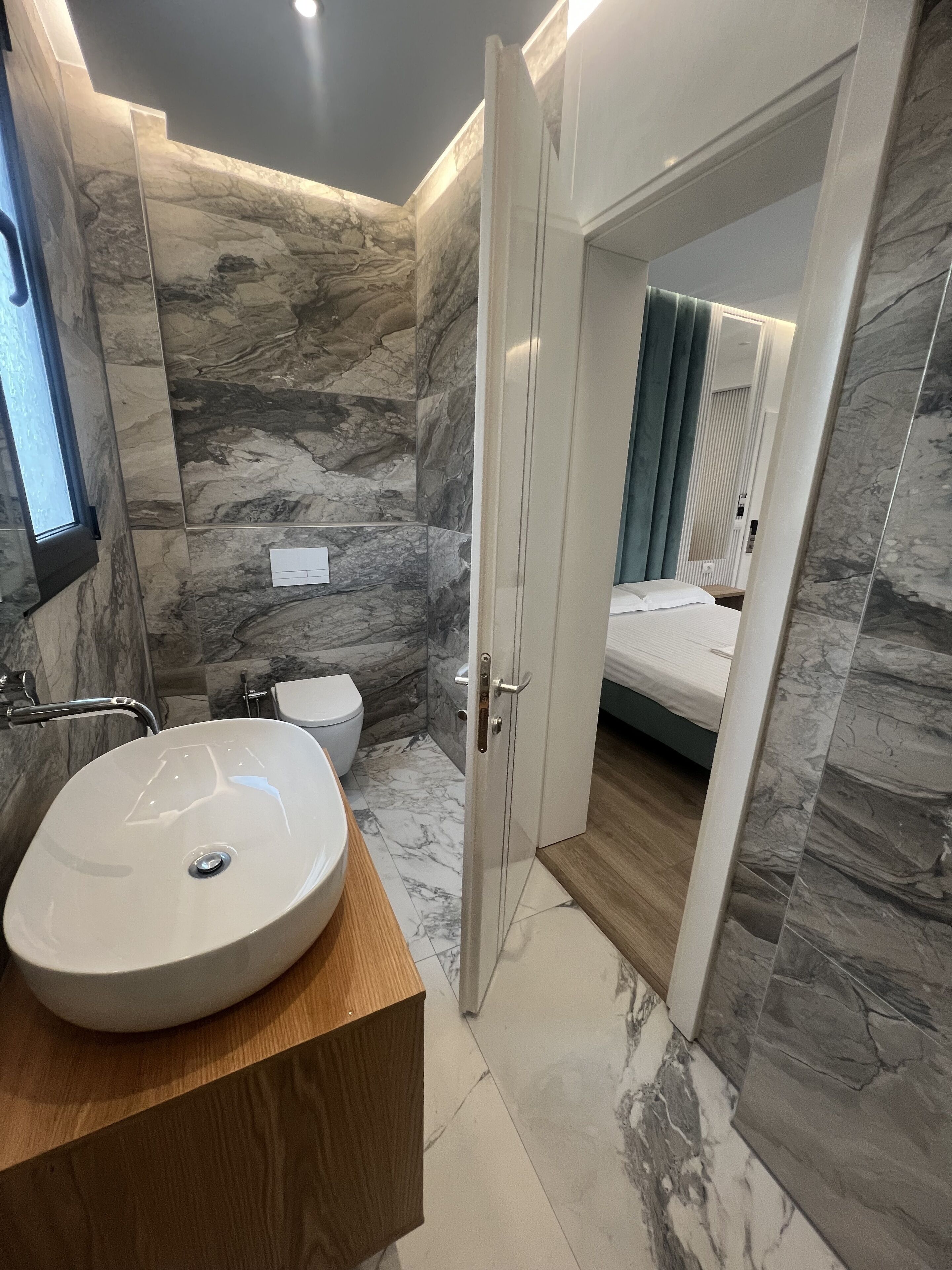 A modern bathroom with a marble wall, a wooden vanity, a white sink, and a toilet.