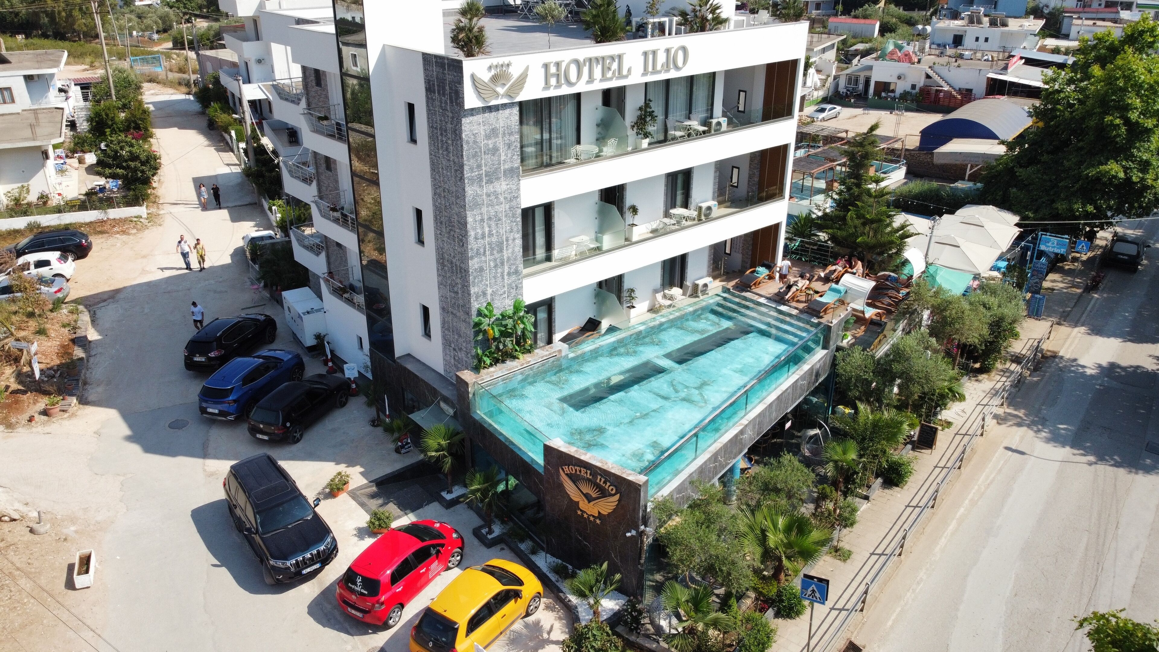 Un hotel moderno con piscina en la azotea, varios balcones y un estacionamiento con varios coches.